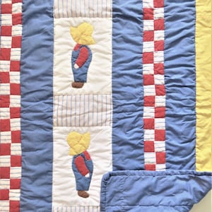May include: A handmade quilt with a patchwork design. The quilt features a repeating pattern of a child figure in overalls, alternating with blue and red checkered squares. The color palette includes blue, red, yellow, and white.