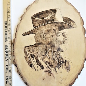 May include: A woodburned portrait of a man wearing a cowboy hat on a wooden slice. The man is facing to the right and has a mustache. The wood slice is approximately 12 inches in diameter.
