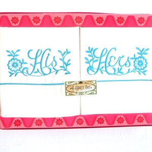 Puede incluir: Un juego de dos fundas de almohada blancas con bordados azules. Las fundas de almohada están en una caja rosa con una etiqueta blanca que dice "Pillow Cases". El bordado en las fundas de almohada dice "His" y "Hers".
