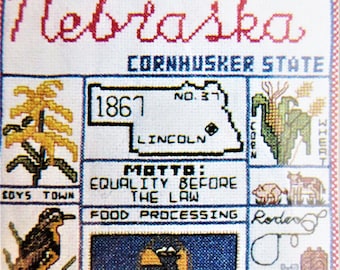 May include: A cross-stitch sampler featuring the state of Nebraska. The sampler includes the state's name, nickname, motto, and a map of the state. The sampler also includes images of corn, a bird, a cowboy boot, and a lasso.