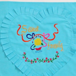 Puede incluir: Una funda de almohada azul claro con un diseño en forma de corazón bordado con las palabras "Sweet Family" en dorado y cintas de colores. El corazón está rodeado por un ribete de volantes.
