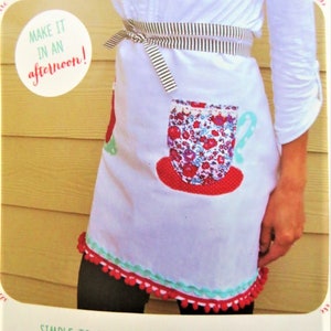 May include: White tea towel apron with a floral teacup design, red pom-pom trim, and a striped waistband. The apron has the text "TEA TOWEL APRON" and "MAKE IT IN AN afternoon!" printed on it.