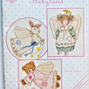 Peut inclure: Un livre de patrons de point de croix intitulé "Priscilla's Babyland", avec des créations de Gloria & Pat et Priscilla Hillman. La couverture présente trois motifs de point de croix : un lapin ange, une fille ange et un ours en peluche ange. Le livre est orné de petits cœurs bleus.