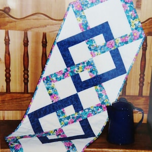 Peut inclure: Chemin de table matelassé avec un motif géométrique. Le chemin de table présente des carrés blancs encadrés de rectangles bleus, avec une bordure florale de fleurs roses, jaunes et bleues. Une cafetière en émail bleu est à proximité.