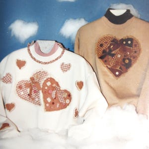 May include: Two sweaters with heart appliqués. One is white with red and pink hearts, the other is tan with a brown heart. The hearts are made of various fabrics and embellished with buttons and bows. The sweaters are displayed against a blue sky background with fluffy white clouds.