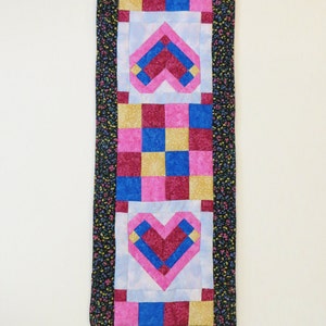May include: A quilted table runner with a black background and a floral pattern. The runner features three pink, blue, and burgundy hearts in the center. The hearts are surrounded by a patchwork of squares in pink, blue, burgundy, and gold.