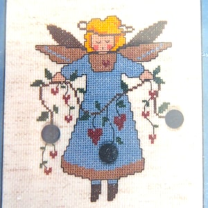 May include: A cross-stitch pattern featuring a blue-dressed angel with blonde hair and wings, holding a vine with red hearts. The angel is standing on a beige background.