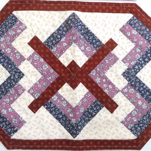 May include: A quilted table runner with a geometric design. The runner features a cream-coloured background with a repeating pattern, accented by burgundy, navy, and pink floral fabric. The edges are trimmed with a burgundy border.