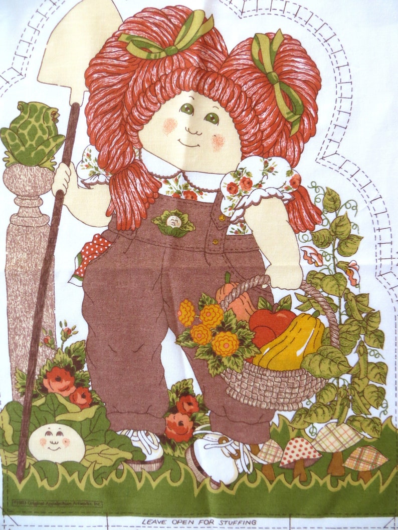 Puede incluir: Una mu&ntilde;eca de pelo rojo con un lazo verde en el pelo sostiene una pala y una cesta de verduras. Lleva un mono marr&oacute;n y una camisa blanca con estampado floral. La mu&ntilde;eca est&aacute; de pie en un jard&iacute;n con hojas verdes y flores. El texto "Leave Open for Stuffing" est&aacute; impreso en la parte inferior de la imagen.