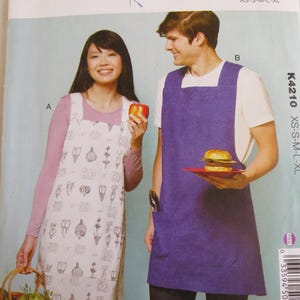May include: Apron sewing pattern by Kwik Sew K4210. The pattern includes two aprons, one with a white background and a floral design, and the other is a solid purple color. The pattern is for sizes XS-XL.