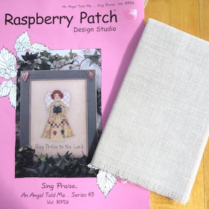May include: A pink Raspberry Patch Design Studio pattern book with an angel cross-stitch design, and a beige linen fabric. The book features the text "Sing Praise" and "An Angel Told Me... Series #3". The fabric is rectangular.