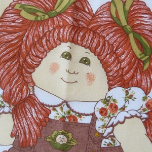 Puede incluir: Una mu&ntilde;eca de pelo rojo con ojos verdes y un vestido de peto marr&oacute;n. La mu&ntilde;eca sostiene una tela blanca con flores rojas y verdes.