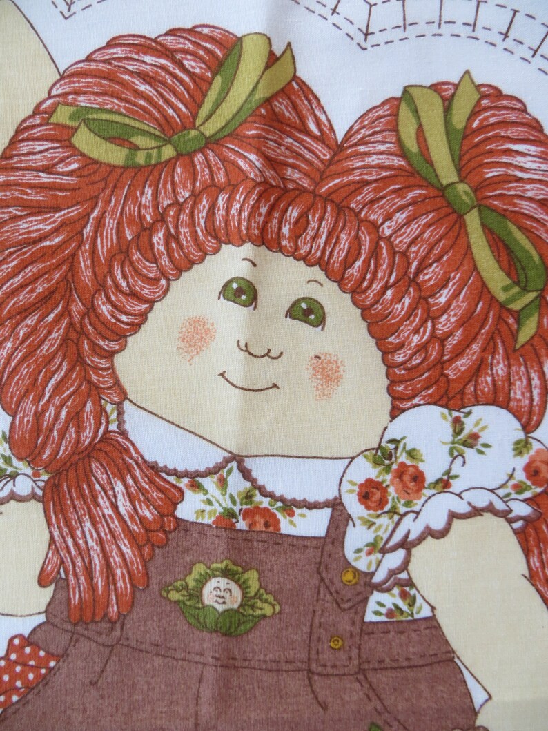 Puede incluir: Una mu&ntilde;eca de pelo rojo con ojos verdes y un vestido de peto marr&oacute;n. La mu&ntilde;eca sostiene una tela blanca con flores rojas y verdes.