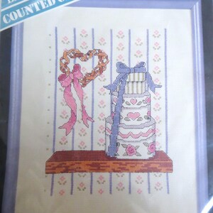 May include: A counted cross stitch pattern featuring a stack of gift boxes with a pink and white heart design, a heart-shaped wreath with pink ribbon, and a wooden shelf. The pattern is on a white background with a light blue and pink striped pattern.