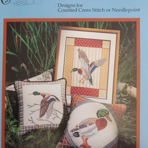 May include: The image showcases a framed cross-stitch design of a mallard duck in flight, alongside two decorative pillows with similar duck motifs. The pillows and framed art feature a mix of colors, including brown, green, and blue, set against a natural backdrop.