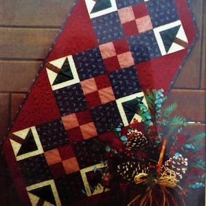 May include: A red, white, blue and green quilted table runner with a geometric pattern. The runner is made of fabric squares and triangles. The runner is displayed on a wooden surface with a pine cone and greenery.