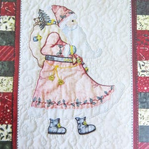 May include: A quilted wall hanging featuring a pink and white embroidered Santa Claus carrying a bag of gifts. The background is a patchwork of red, green, and white fabric.