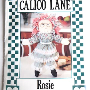May include: A Simplicity sewing pattern titled "Calico Lane" featuring a doll named "Rosie." The doll is dressed in a floral print dress with lace trim, pink hair, and glasses. The pattern cover has a white background with green and white checkered borders.