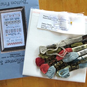 May include: A cross stitch kit with a white fabric, a printed pattern, and a variety of embroidery floss in shades of red, blue, green, and beige. The pattern is titled "Button Friends" and includes the text "Friends are like a collection of buttons, each unique in size, shape and color."