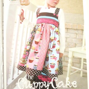May include: A young girl wearing a colorful cupcake-themed dress. The dress features a white top, brown and pink accents, and a skirt with cupcake patterns in pink, blue, and brown. The text "the Cuppy Cake dress" is visible.