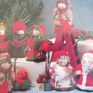 May include: A set of eight handmade Christmas ornaments featuring a variety of characters, including a Santa Claus, a baby Jesus, and several carolers. The ornaments are made from felt and fabric and are decorated with beads, sequins, and other embellishments.