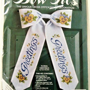 Puede incluir: Un kit de punto de cruz para una decoración de puerta en forma de lazo. El kit incluye una tela blanca con un diseño floral y las palabras "Greetings" bordadas en ella. El kit también incluye hilo, una aguja e instrucciones.