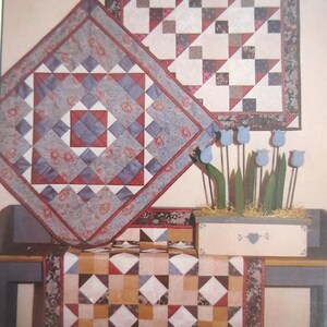 May include: Three quilted wall hangings with geometric patterns in shades of red, blue, white, and brown. The hangings are displayed on a wall with a wooden box filled with straw and artificial flowers.