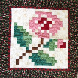 May include: A white quilt with a pink and green pixelated flower design. The quilt is surrounded by a black and red floral border.