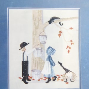 May include: Cross-stitch pattern featuring two figures in traditional clothing standing near a tree with a bucket hanging from a branch. A black cat sits on the branch above them. A goose stands to the right of the figures. The background is a white fabric with red and brown leaves scattered around the figures.