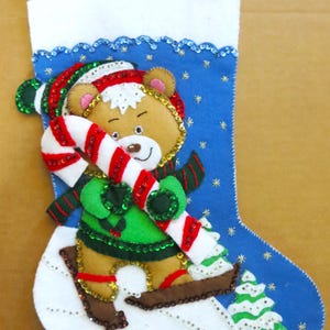 Könnte beinhalten: Blaue Weihnachtsstrumpf mit einem Teddybären aus Filz, der eine Zuckerstange hält. Der Bär trägt ein grünes Outfit, einen rot-grünen Hut und einen Schal. Der Strumpf hat eine weiße Manschette, Schneeflocken-Akzente und einen kleinen Weihnachtsbaum.