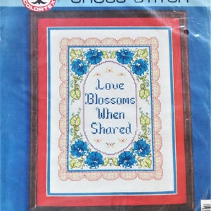May include: Counted cross stitch pattern featuring a white background with blue flowers and green leaves. The text "Love Blossoms When Shared" is stitched in black. The finished size is 9 inches by 12 inches and the matted size is 11 inches by 14 inches.