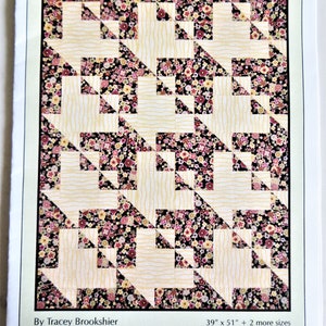 May include: A quilt pattern guide featuring a quilt with a tessellated design of interlocking baskets. The quilt is made with a black and white fabric with a floral pattern and a cream fabric with a wavy pattern. The quilt is 39 inches by 51 inches and comes in two other sizes. The title of the pattern is "Interlocking Baskets" and the subtitle is "Traditionally Pieced Basket Quilt with a Tessellated Twist!" The pattern is by Tracey Brookshier and the pattern number is #07766.