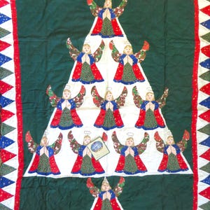 May include: A white quilt with a green border and a Christmas tree design featuring angels. The angels are arranged in a pyramid shape with a star at the top. The quilt is decorated with red, white, and blue triangles.
