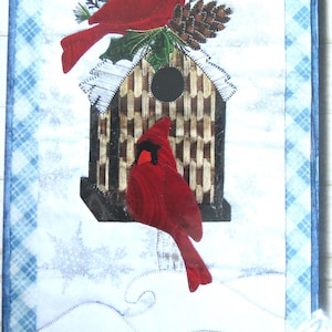 May include: A winter-themed quilted wall hanging featuring two red cardinals perched on a birdhouse with a pine cone on top. The birdhouse is brown and white with a black opening. The background is a blue and white plaid pattern with snowflakes.