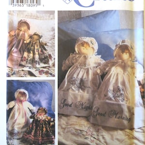 May include: A sewing pattern for two dolls with embroidered details. The dolls are dressed in white dresses with floral embroidery and have the text "Good Night" and "Good Morning" embroidered on their dresses. The pattern is from Simplicity Crafts.