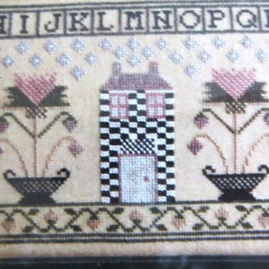 May include: Cross-stitch sampler with a black and white checkered house, two pink flower arrangements, and the alphabet in black letters.
