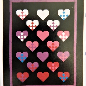 May include: A quilt with a black background and a purple border. The quilt is made up of 19 hearts in various shades of pink, purple, and white. Each heart has a different pattern of squares.