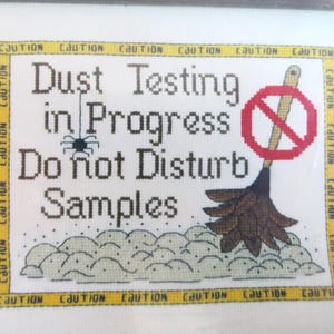 Può includere: Un cartello bianco con bordi di nastro giallo di cautela e testo nero che recita "Dust Testing in Progress Do not Disturb Samples". Un cerchio rosso con una linea rossa diagonale attraverso di esso si trova nell'angolo in alto a destra del cartello. Una scopa marrone con un manico nero si trova sotto il cerchio.