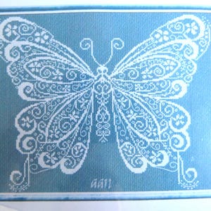 May include: A white butterfly with intricate details is embroidered on a light blue background. The butterfly is centered on the canvas and has a delicate, lacy design. The word "May!" is written in white at the bottom of the canvas.
