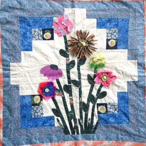 May include: A white quilt with a blue and white border. The center of the quilt features a floral design with pink, purple, blue, and green fabric flowers. The flowers are arranged in a bouquet with green fabric stems.
