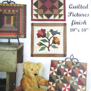 May include: A display of three quilted pictures, each approximately 10 x 10 inches. The quilts feature various geometric and floral patterns in shades of red, brown, and green. A teddy bear sits in front of one of the pictures.