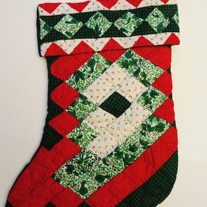 Può includere: Un calzino di Natale trapuntato verde e rosso con un motivo a rombi. Il calzino ha un bordo bianco e verde intorno alla parte superiore.