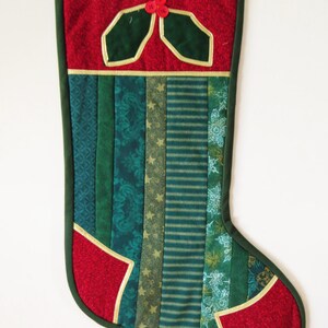 Peut inclure: Une chaussette de Noël rouge et verte avec un motif de houx et une bordure dorée. La chaussette est faite de tissu patchwork et a une boucle en haut pour l'accrocher.