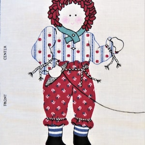 Op de afbeelding: Een roodharige pop met een blauw-wit gestreept shirt, rode broek met een wit stippenpatroon en zwarte laarzen. De pop houdt een touw vast in haar rechterhand en heeft een rode appel op haar shirt.