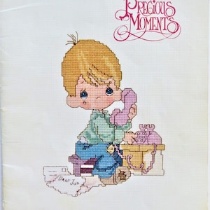 May include: A cross-stitch pattern featuring a young boy with blonde hair, blue eyes, and a green shirt, sitting on a stool and talking on a pink phone. The text "Dear Jon" is written on a piece of paper in the lower left corner of the image. The pattern is from the Precious Moments collection.