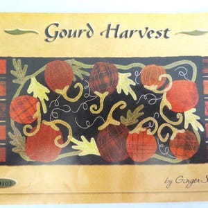 May include: A rectangular craft pattern titled Gourd Harvest by Ginger Sanchez. The design features a black center panel with orange and red plaid gourds, yellow leaves, and swirling vines. The edges have orange plaid tabs on both sides.