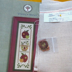 May include: A cross stitch kit for a project called "Granny's Apples". The kit includes a piece of fabric, floss, and a pattern. The fabric is a light beige linen with a red border. The floss is in a small plastic bag. The pattern is printed on a piece of paper with a red border. The pattern is for a cross stitch design of three apples in a bowl.