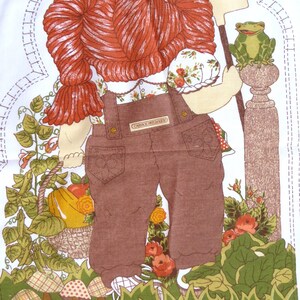 Puede incluir: Una mu&ntilde;eca de tela marr&oacute;n y blanca con pelo rojo que lleva un mono y una camisa blanca con estampado floral. La mu&ntilde;eca sostiene una pala y est&aacute; de pie junto a una rana verde sobre un pilar marr&oacute;n. La mu&ntilde;eca est&aacute; rodeada de hojas verdes y flores.