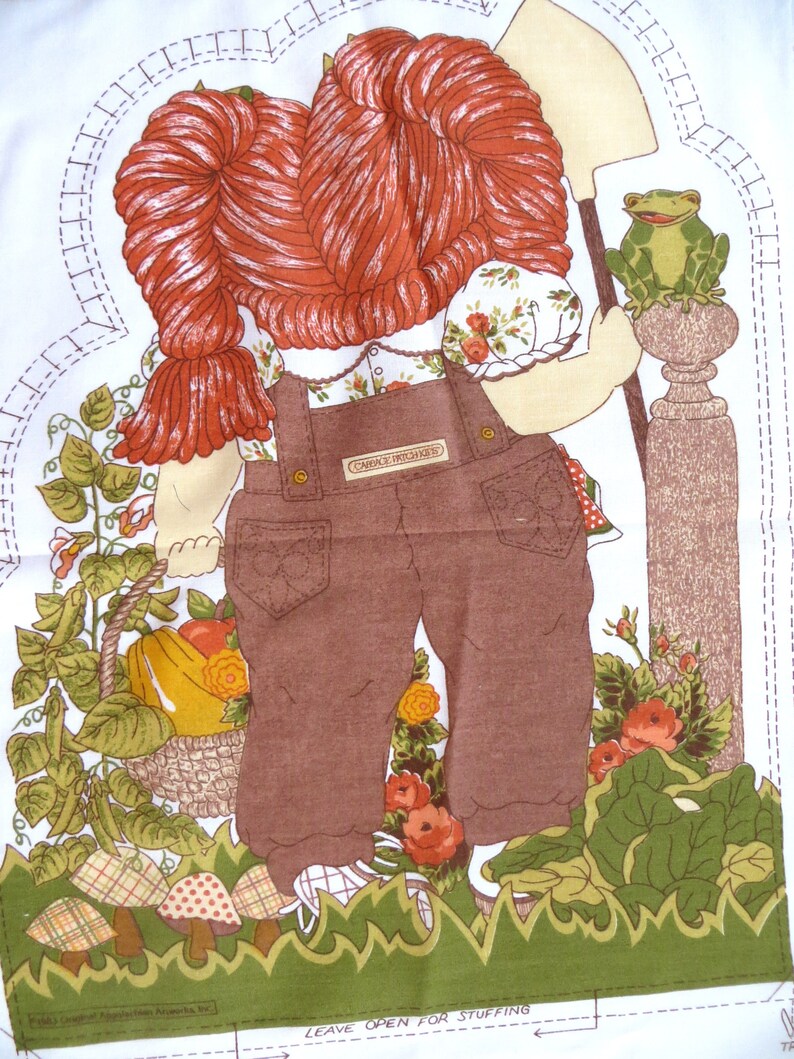 Puede incluir: Una mu&ntilde;eca de tela marr&oacute;n y blanca con pelo rojo que lleva un mono y una camisa blanca con estampado floral. La mu&ntilde;eca sostiene una pala y est&aacute; de pie junto a una rana verde sobre un pilar marr&oacute;n. La mu&ntilde;eca est&aacute; rodeada de hojas verdes y flores.