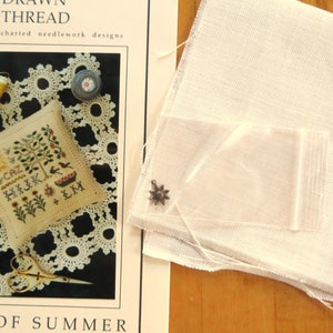 May include: A cross stitch pattern for a pillow featuring a tree, flowers, and the text "Pot of Summer". The pattern is on a white background with a black border. The image also includes a piece of white fabric, a small bag, and a metal charm.